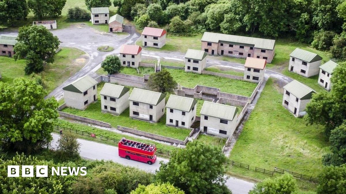 An aerial picture of what the village looks like today, with a small cluster of empty brick houses with tin roofs and no windows or doors. It is a stark and bare street and all the houses look identical. At the bottom there is a red double-decker tour bus driving along the road.