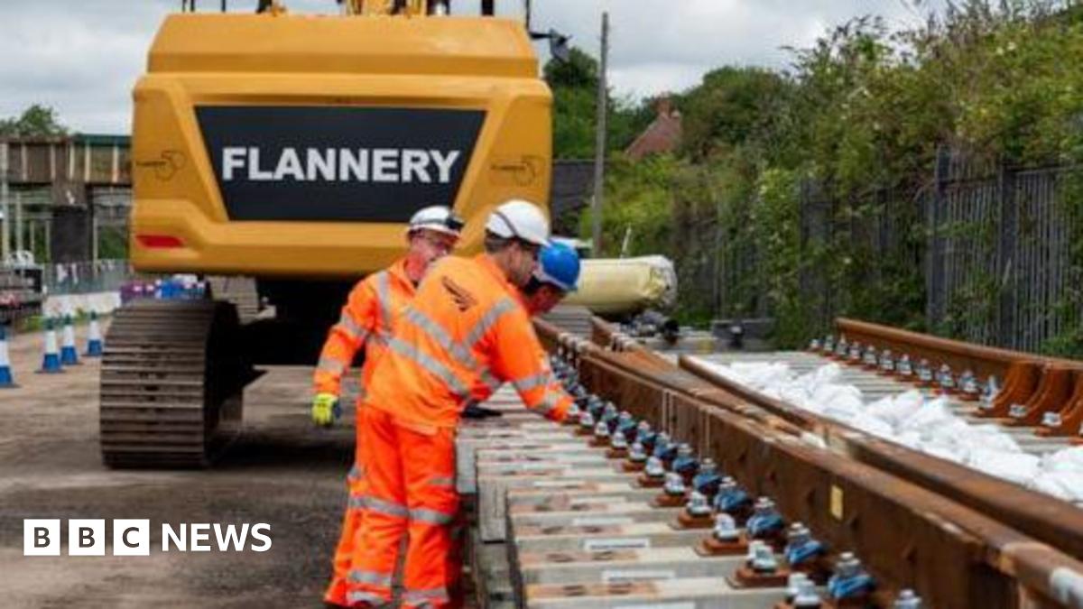 Network Rail employees dressed in orange overalls, wearing white and blue hats inspect new tracks before being lifted into position by a digger.