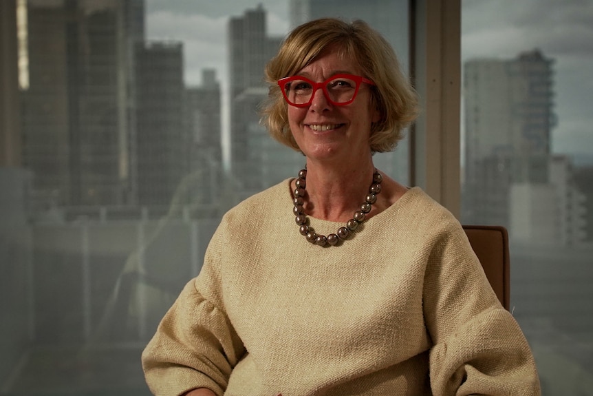 Woman with mousy blonde hair and wearing red-framed glasses smiling while seated