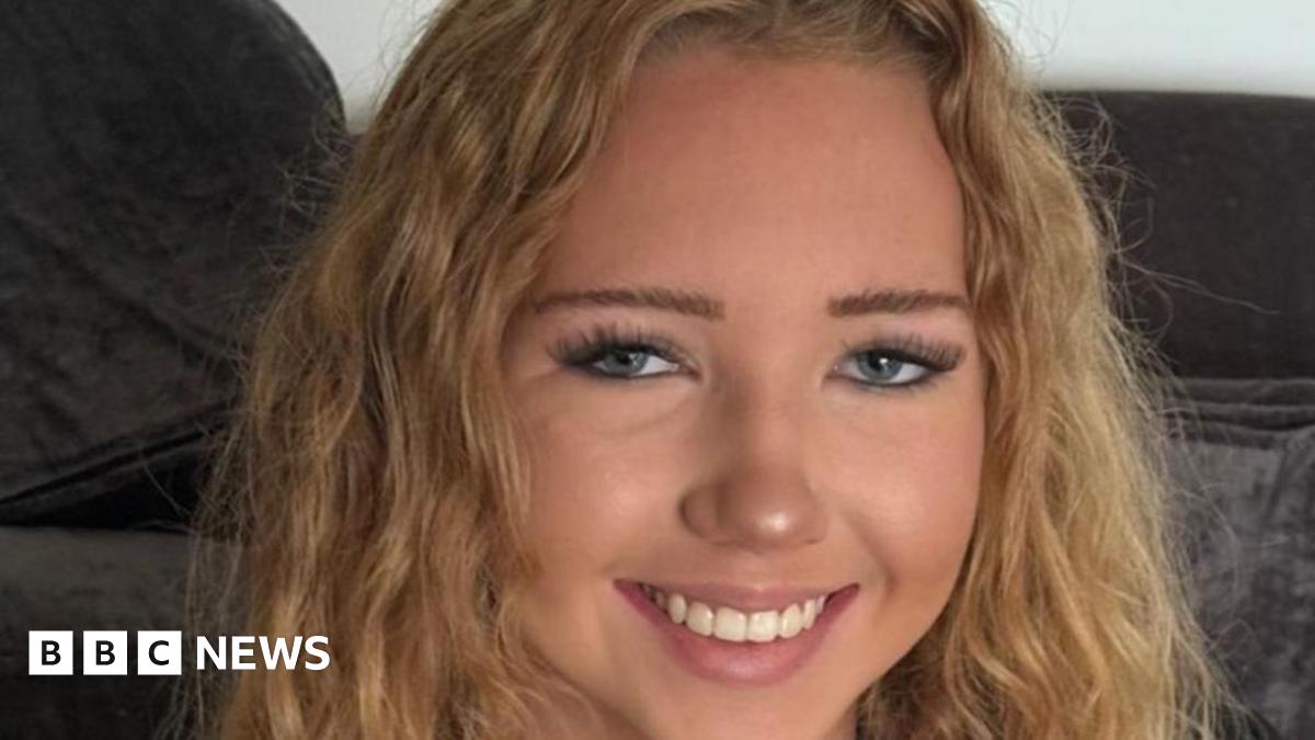 A young girl with curly blonde hair, she is wearing mascara and has long eyelashes. She is smiling at the camera