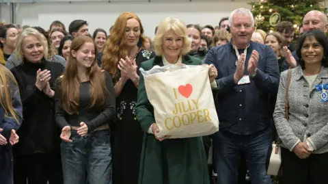Reuters Britain's Queen Camilla holds a tote bag with the name of Jilly Cooper on it, who wrote the original books entitled Rivals, as she poses with members of the cast and crew during a visit to the set of the TV series Rivals.