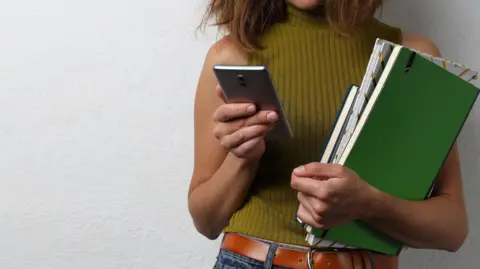 Getty Images A stock image shows a woman from the chin down wearing a yellow sleeveless top and jeans. She is holding several folders and notepads under her left arm and a phone in her right hand, which she appears to be locking at. She is standing in front of a white textured wall.