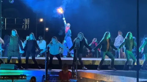 Arzu Dutta/BBC Female performers form a circle holding hands on stage. In the middle of them is another performer holding a fire torch above her head.