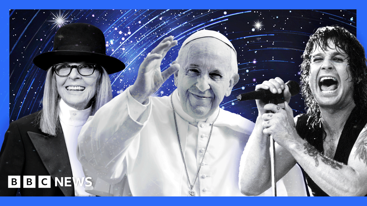 D’Angelo is performing on stage, wearing a wide-brimmed black hat and a black leather vest over a deep V-neck top. A silver chain necklace is visible around the neck, and the left arm shows a detailed tattoo design. He is positioned near a microphone with a black foam cover. The background features a warm orange gradient, creating a dramatic and vibrant concert atmosphere.