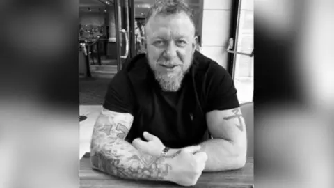 Facebook A photograph taken from Facebook of Mark Killick, who is leaning on a table top. He is wearing a black short-sleeved top and has extensive tattoos on his arms, which are very muscled. He has a goatee beard and receding hair