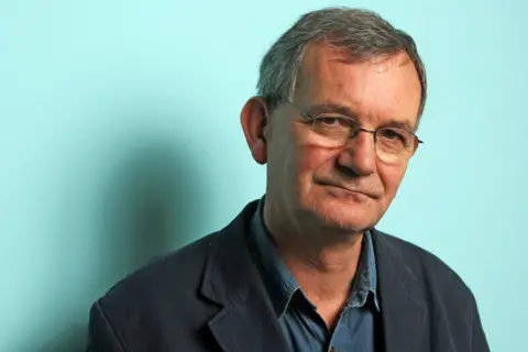 Getty Images Martin Parr is wearing a dark collared shirt layered with a black blazer, standing against a plain light teal background. The lighting creates a soft shadow on the wall behind, adding depth to the otherwise simple setting. The overall composition suggests a formal or professional portrait style.