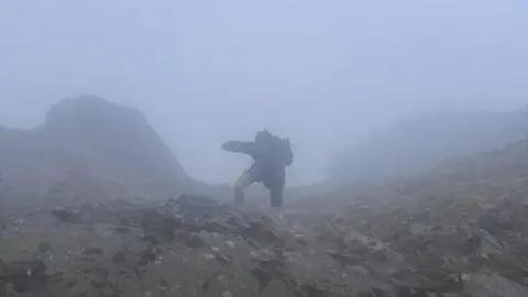 Nathan Mburu A man standing on a ridge of a mountain. He is mid climb and holding on to the ground in front of him. The weather conditions around him are foggy. 