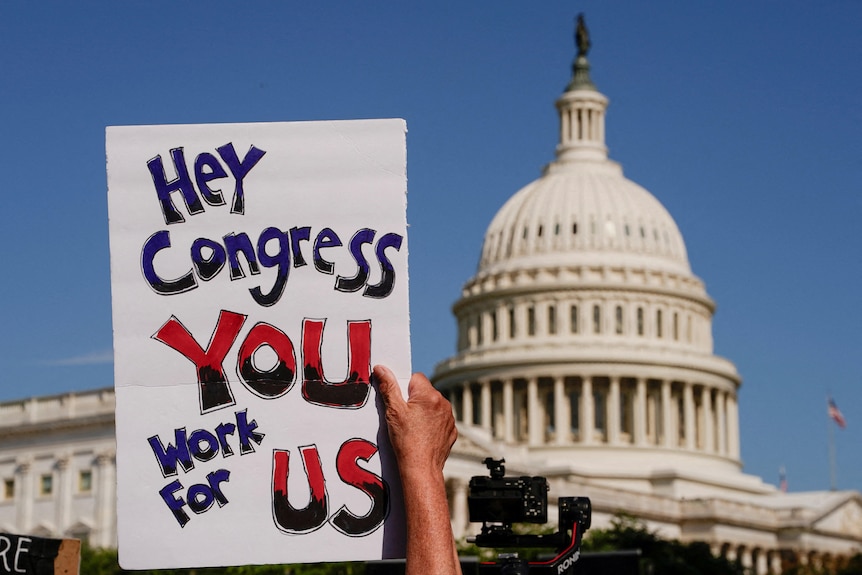 A person holding up a sign saying 'Hey Congress, You Work For Us', with the US Capitol in the background.