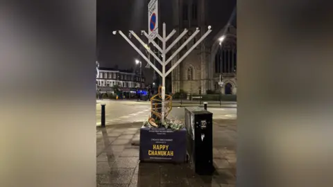 Jason Groves A large decorative silver Hanukkah menorah on a street, on a stand which reads 'Happy Chanukah'. The lightbulbs representing candles on the top of the menorah have been damaged.