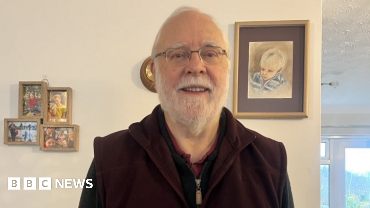 A man with short grey hair and a grey beard, wearing glasses. He has a dark zipped top, and is smiling at the camera, stood in front of a wall inside a house.
