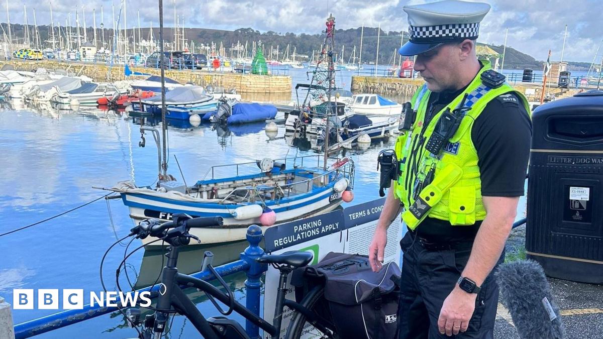 A police officer in a bright yellow vest stands in front of a bike leaned next to a railing. In the background there are boats in a harbour.