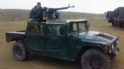 Lasha Shergelashvili Lasha wearing army fatigues and a cap standing on an armoured vehicle and resting his arm on a mounted rifle 