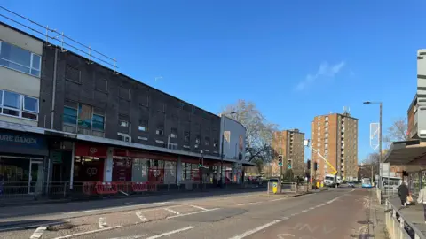 Bedford Borough Council A road with a three-storey shop and living space development to the left and right, and tower blocks in the background. There is also a tree between the flats and the tower blocks. It is sunny and there is no traffic on the road.