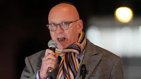 PA Media Lord Wolfson speaking into a microphone. He is a bald man wearing glasses and a striped scarf with a grey coat.