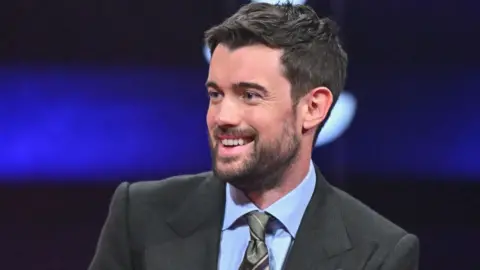 PA Jack Whitehall wearing a suit and grinning. He has dark hair and a beard.