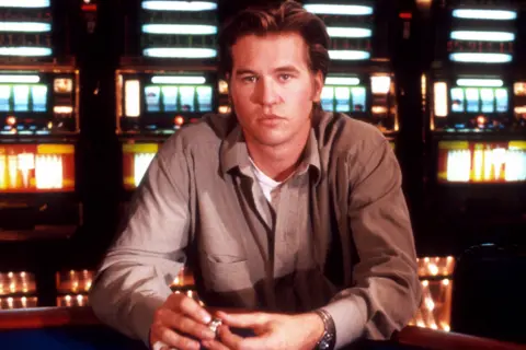Getty Images Val Kilmer is seated at a casino table with hands resting on the edge, holding a small object. He is wearing a light-coloured button-up shirt over a white undershirt and a wristwatch. In the background, there are several brightly lit slot machines arranged in rows, creating a colourful and vibrant setting typical of a casino environment. The photo is a still from his 1989 film, Kill Me Again.