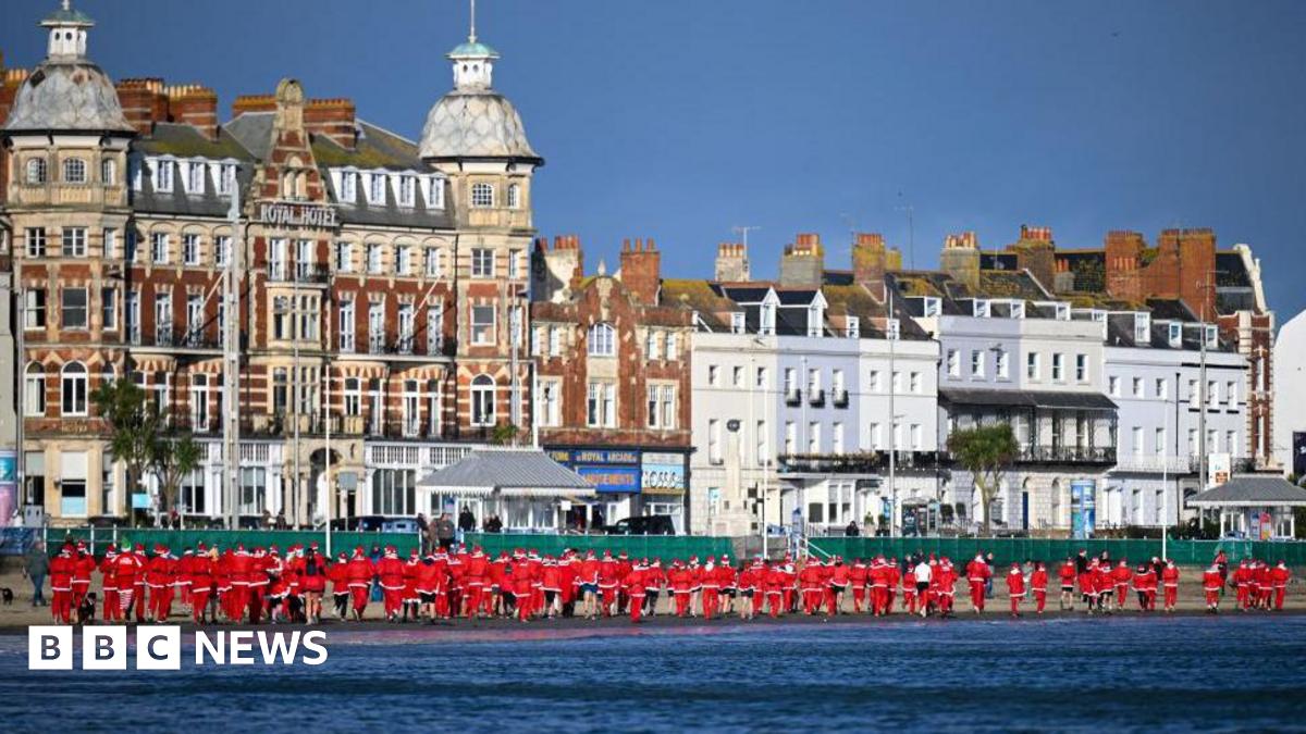Line of runners in red Santa outfits making their way along Weymouth Beach with the sea to the right and buildings along the left.