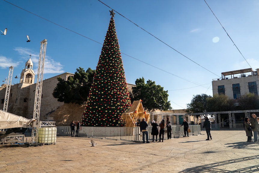 A large christmas tree with a big star in front in the middle of a square in front of a church, with people walking around it.