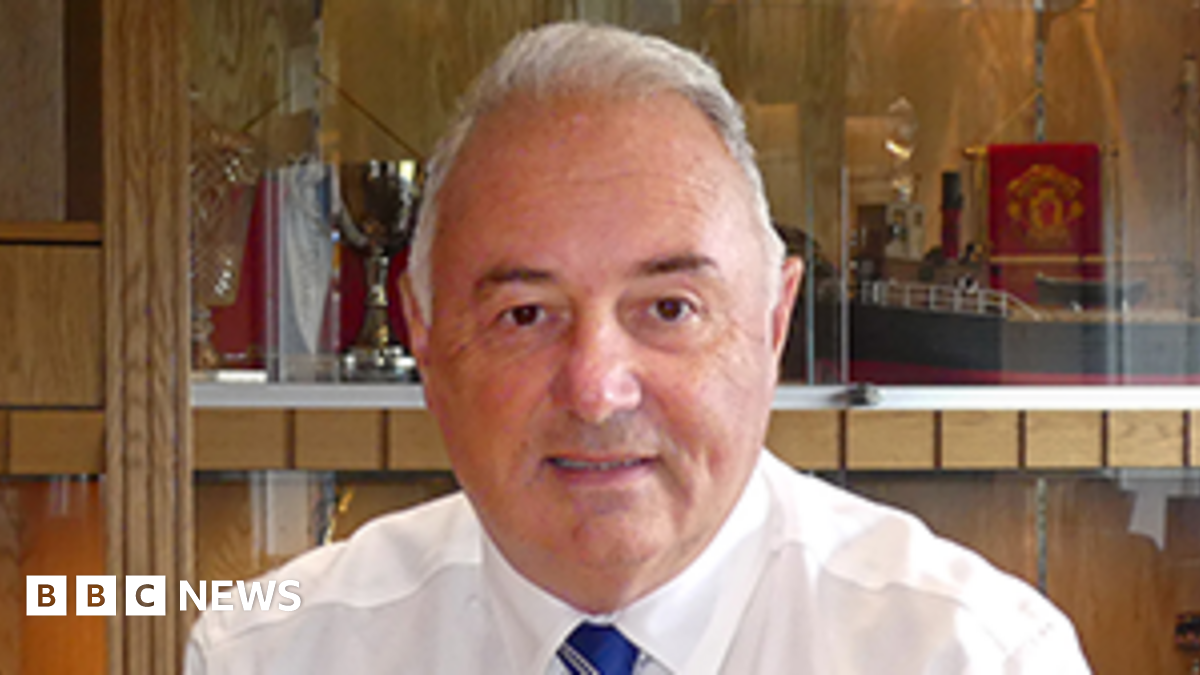 John Laird sitting behind a desk in a trophy room. He has short white hair, and is wearing a white shirt and blue tie.