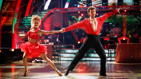 BBC George Clarke and Alexis Warr on the Strictly dancefloor in red samba-style clothes. The stage is lit up with bright lights, with stairs at the back of the ballroom floor. Restaurant-style tables with gingham cloths and black chairs are set up around the stage. No-one is sitting at them.