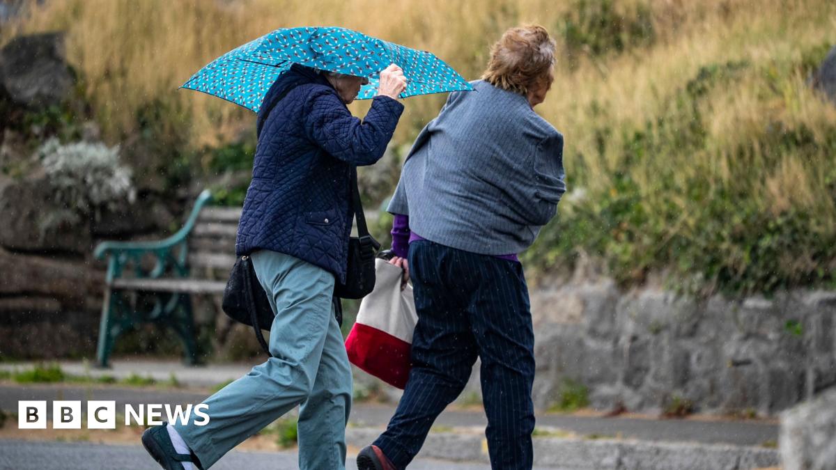 Umbrellas are out as two people are walking along a path. They are both wearing jackets and there is a bench pictured in front of them.