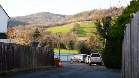A small residential road with fences and cars running alongside. In the background is rolling countryside hills lit up by the sun.