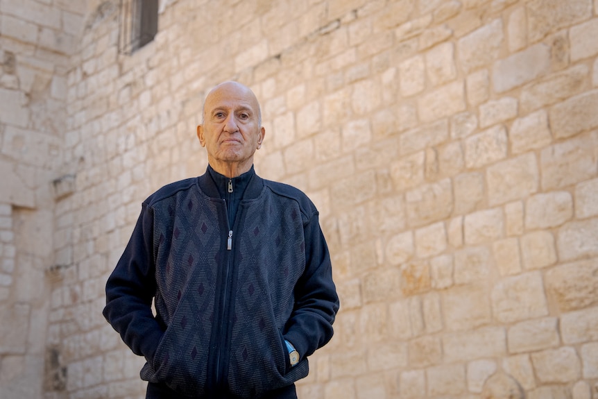An older man, wearing long sleeved jumper, has his hands in its pockets, and standing in front of a wall.