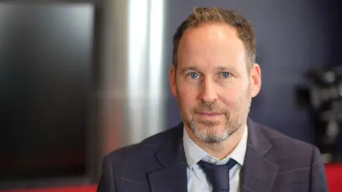 Jamie Niblock/BBC Detective Inspector Yoni Adler is wearing a navy suit jacket, navy tie and pale blue shirt. He has short brown hair and greying stubble.