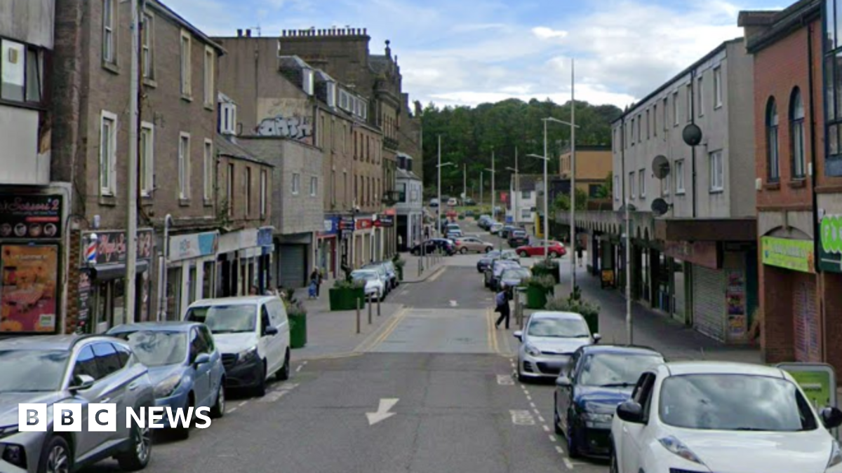 A high street with shops on each side. Cars are parked on either side of the road.