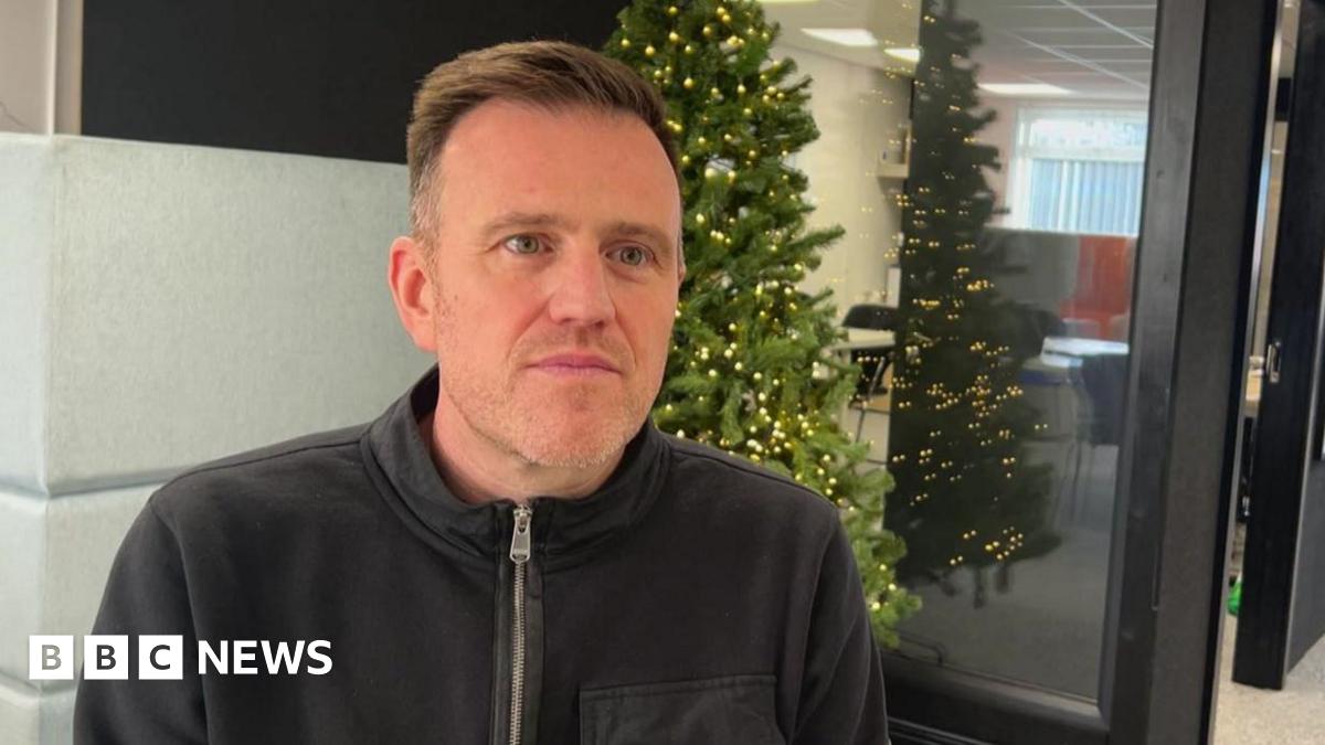 A man wearing a t-shirt which says 'The Place' on it smiles at the camera. Next to him another man wearing glasses and a blue shirt and jacket is looking at the camera. There are posters behind them in a room with white walls.