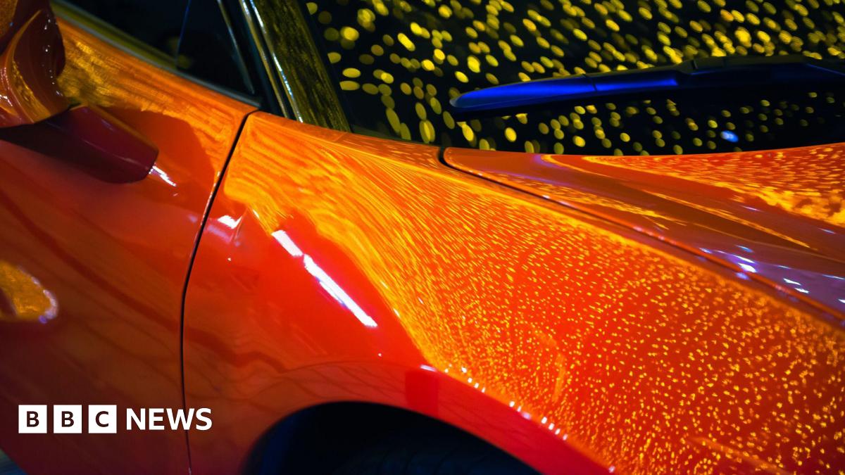 A car, showing part of its front, a door, the colour looks orange with reflections on it.