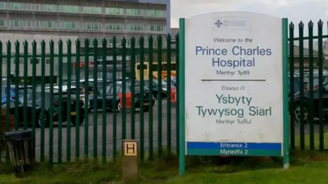 Getty Images A sign outside Prince Charles Hospital, in Merthyr Tydfil. A car park and a large, grey concrete building can be seen behind a metal green fence.