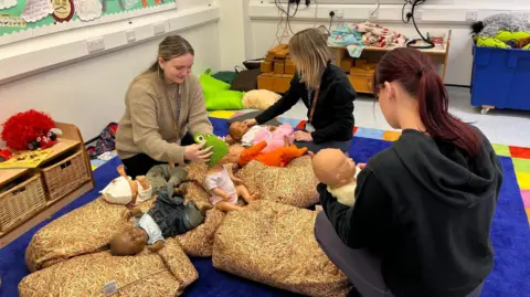 BBC Three female students each caring for a toy doll in a nursery play area setting. There is a screen on in the background playing a video of a working nursery.
