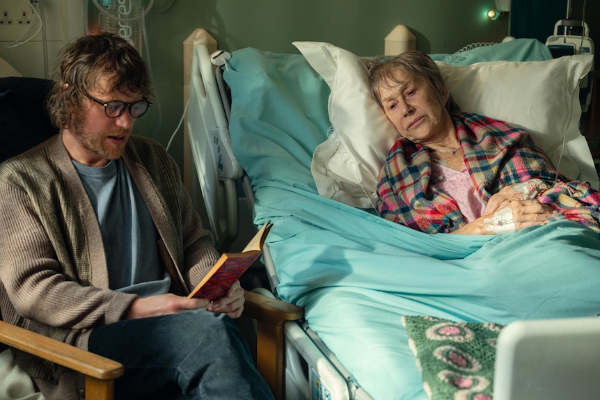 A bearded and bespectacled man sits reading a book to a woman in a hospital bed.