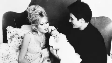 Keystone-France/Gamma-Keystone via Getty Images Brigitte Bardot sits in bed, wearing a light-coloured nightie, her blonde hair half piled on her head, wrapped in floral bedsheets, while Jacques Charrier, wearing a dark shirt and trousers, holds their newborn baby Nicolas in a white outfit.