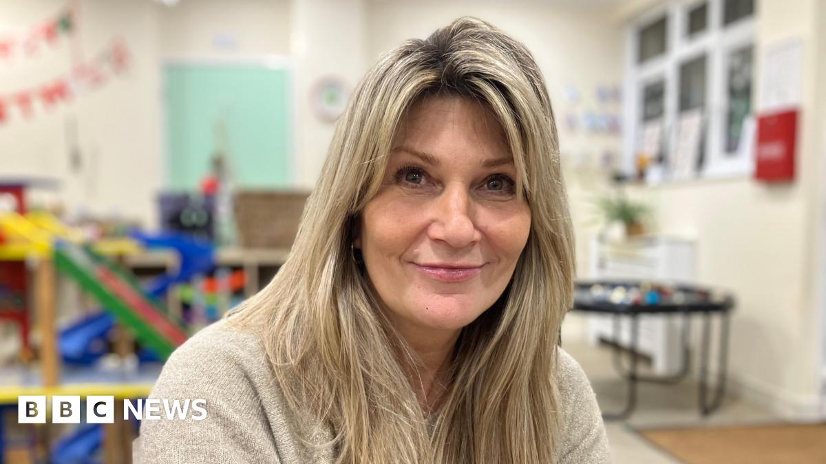 A head and shoulders shot of a woman with long blonde hair, wearing a grey jumper, sitting in a classroom