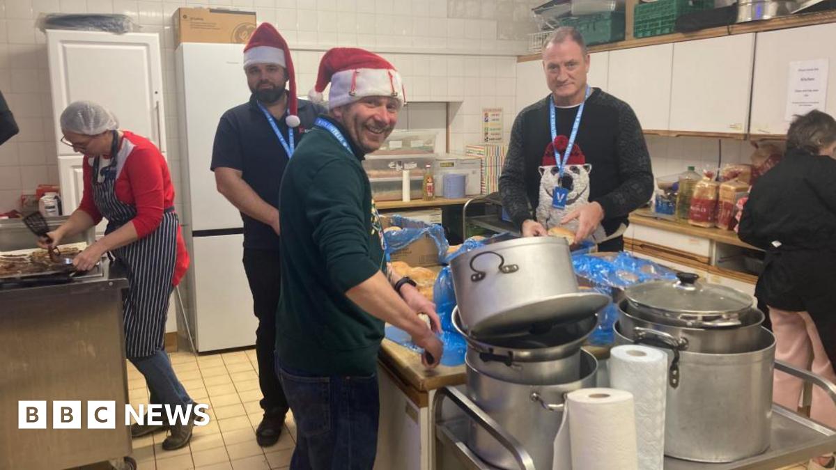 Three men in a kitchen preparing food. Two of them are wearing Christmas hats. A woman is to the left preparing food on a different worktop, as well as another woman on the right. There are metal pots and pans piled up in front of the worktop in the middle.
