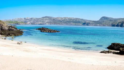 Getty Images A sandy beach on Handa on a beautiful sunny day. The shallow sea water reflects the blue of the sky above. On the opposite shore are low rocky hills.