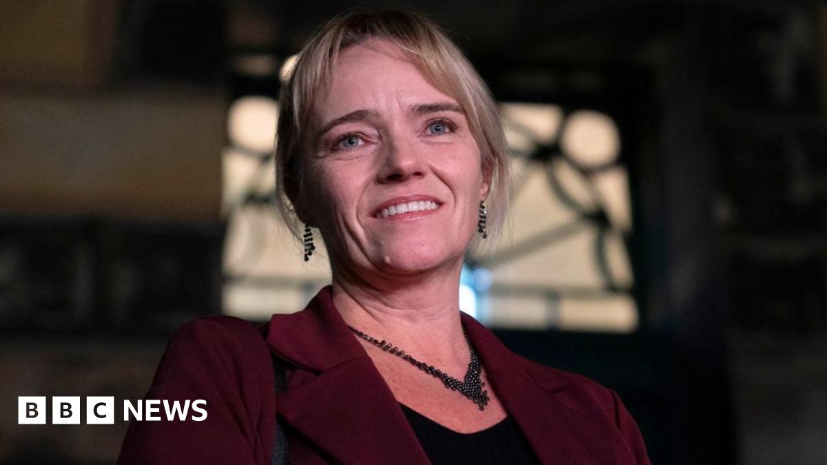 A portrait of a woman, shown from the shoulders up, with a dark background and soft lighting. She has medium-length blonde hair that is neatly styled.