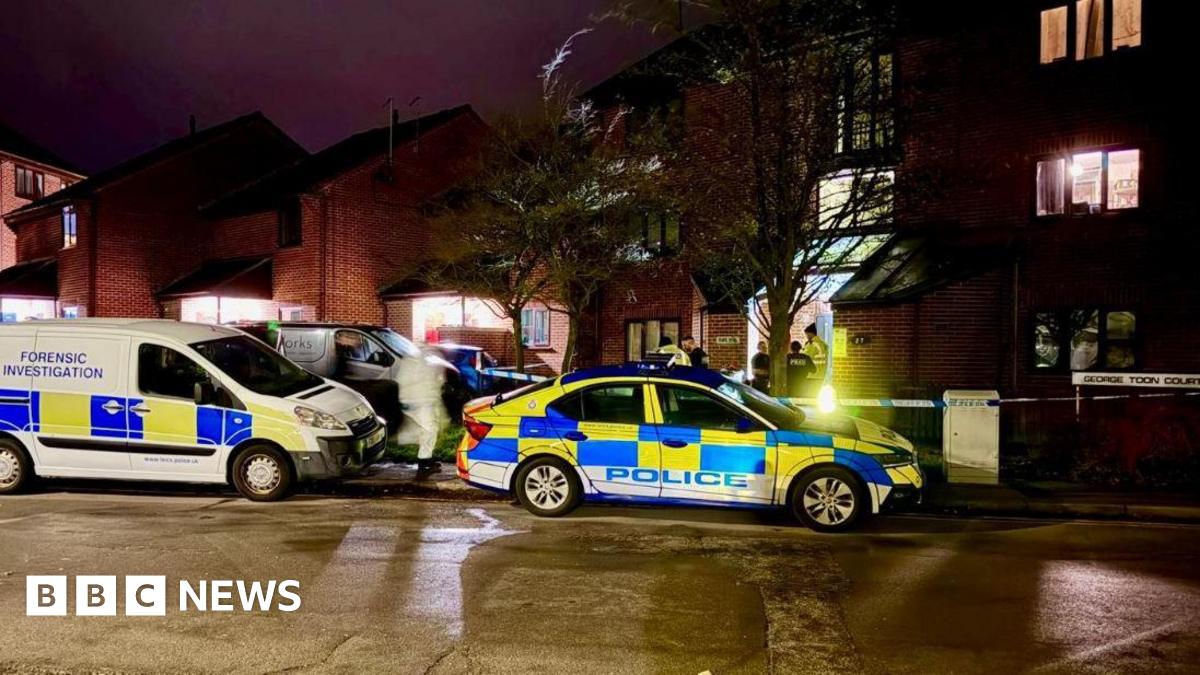 A number of police vehicles outside a block of flats at night. One van says FORENSIC INVESTIGATION. A forensic officer in white overalls is visible from the road. There is also police tape across the entrance way to the block.