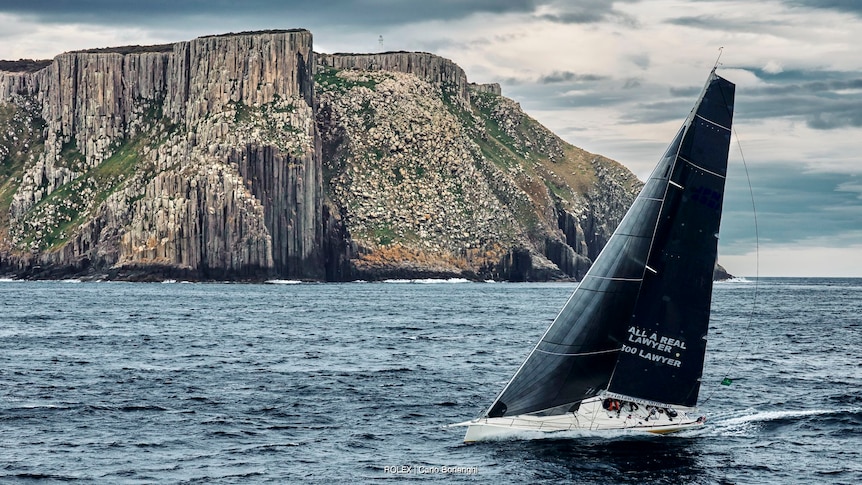 Yacht sailing past a large rocky coastline.