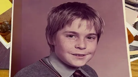 Family photo An old school photograph showing a young boy with freckles. He is wearing a grey shirt and black and gold tie under a great v-neck jumper. 