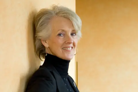 Getty Images Joanna Trollope has short, light-coloured hair and is leaning against a textured beige wall. The individual is wearing a black turtleneck sweater layered with a dark blazer. A small decorative brooch is pinned to the blazer, and a single drop earring is visible. The background is plain, creating a simple and elegant setting.