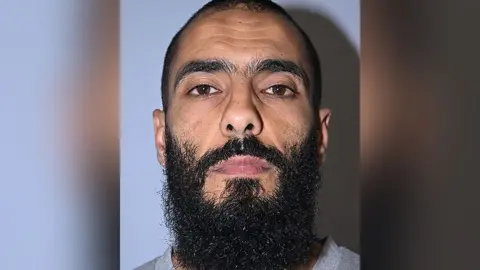 GMP Police custody image of Bilel Saadaoui. He has short black hair and a long black beard and is wearing a grey top.