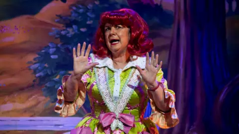 King's Theatre Elaine C Smith performing on stage in a pantomime. She is wearing a bright red wig, and a bright green and pink dress. She is holding her hands in the air as she performs. 
