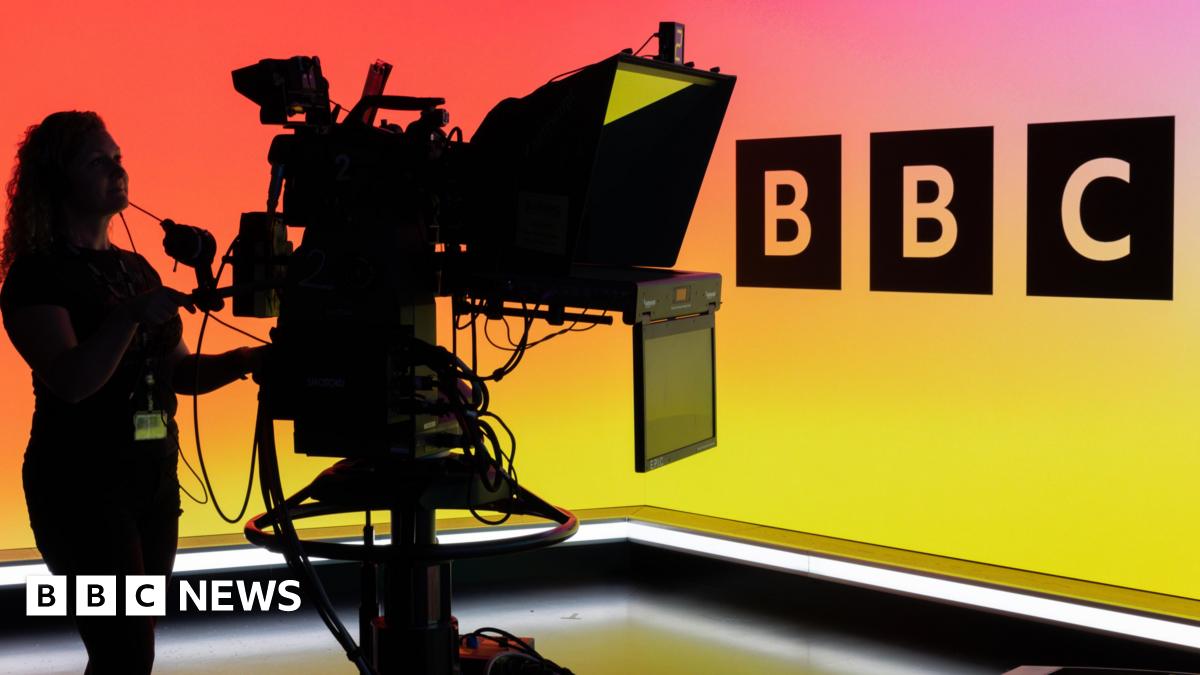 A woman operating a TV camera in front of the BBC Logo on an LED wall.