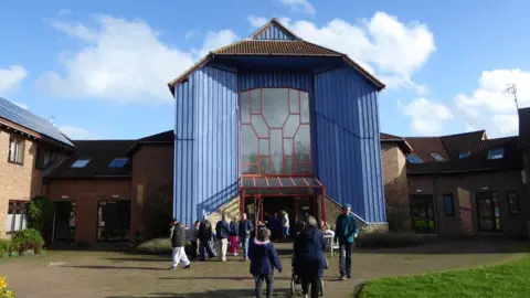 Camphill Milton Keynes A large entrance with blue cladding, a glass front and a pointed roof.