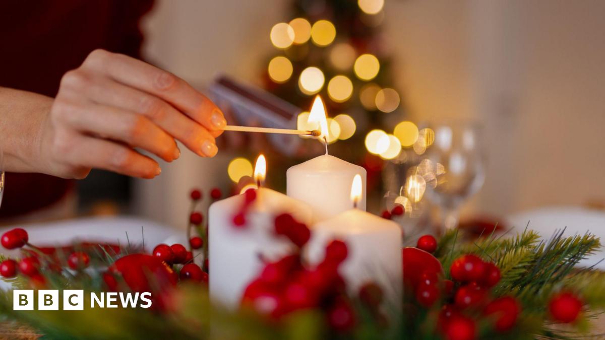 A person lights a white candle with a match on a festive table decorated with holly and red berries, with a softly lit Christmas tree blurred in the background.