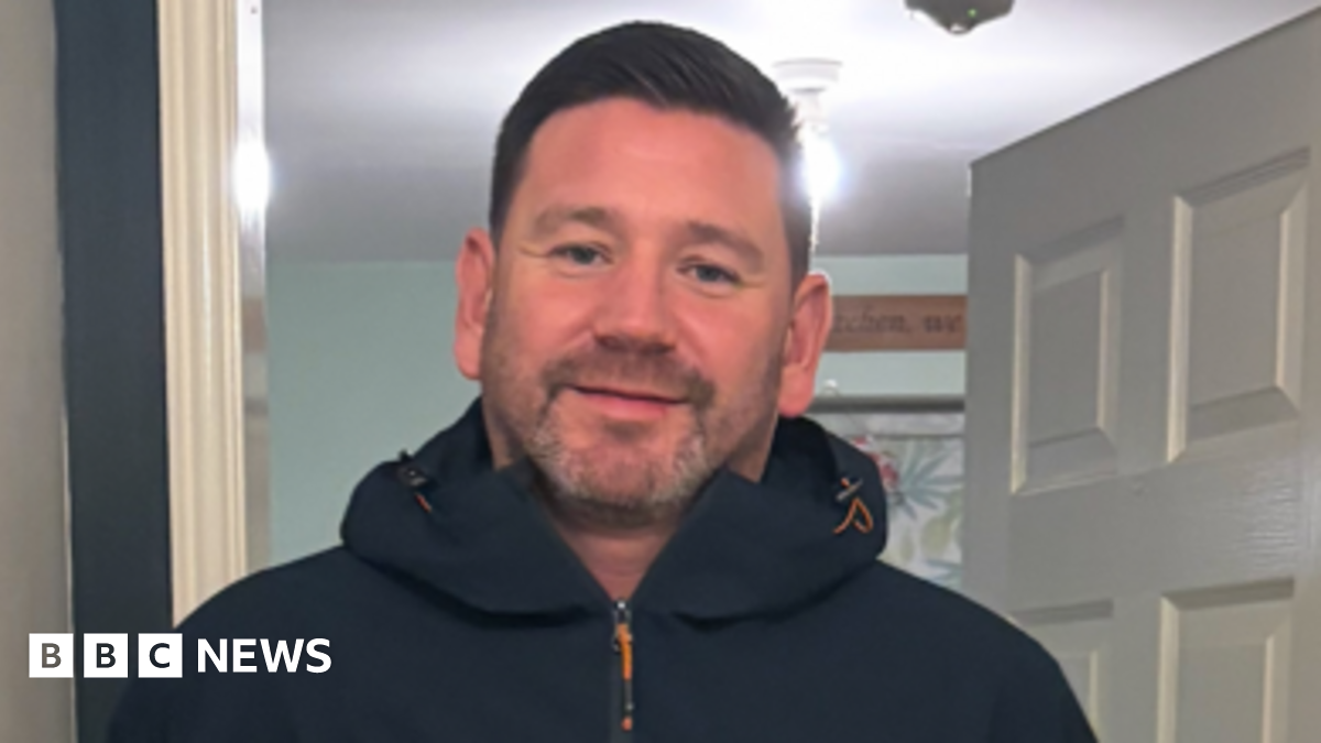 Russell Stamp. He has brown hair and stubble on his chin. He is wearing a hooded coat and is standing in a doorway.