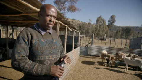 Thabo Makopo is standing by a cattle pen  with some goats visible. He is wearing a patterned jumper and is holding a small water bottle.